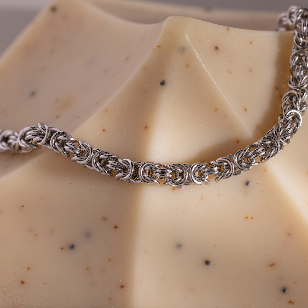 The Bold Byzantine Bracelet, featuring woven silver links, rests on a beige speckled geometric soap bar, radiating quiet luxury.