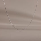 The Dainty Herringbone Necklace in Sterling Silver is displayed on a neutral beige background, ideal for adding a subtle shine to your layered look.