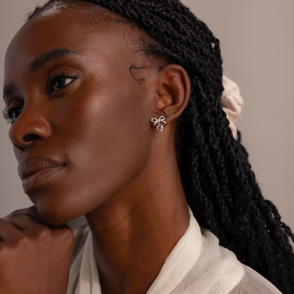 Model wearing delicate silver bow earrings with heart charms, styled with a sheer ivory blouse and soft natural lighting.