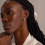Model wearing delicate silver bow earrings with heart charms, styled with a sheer ivory blouse and soft natural lighting.