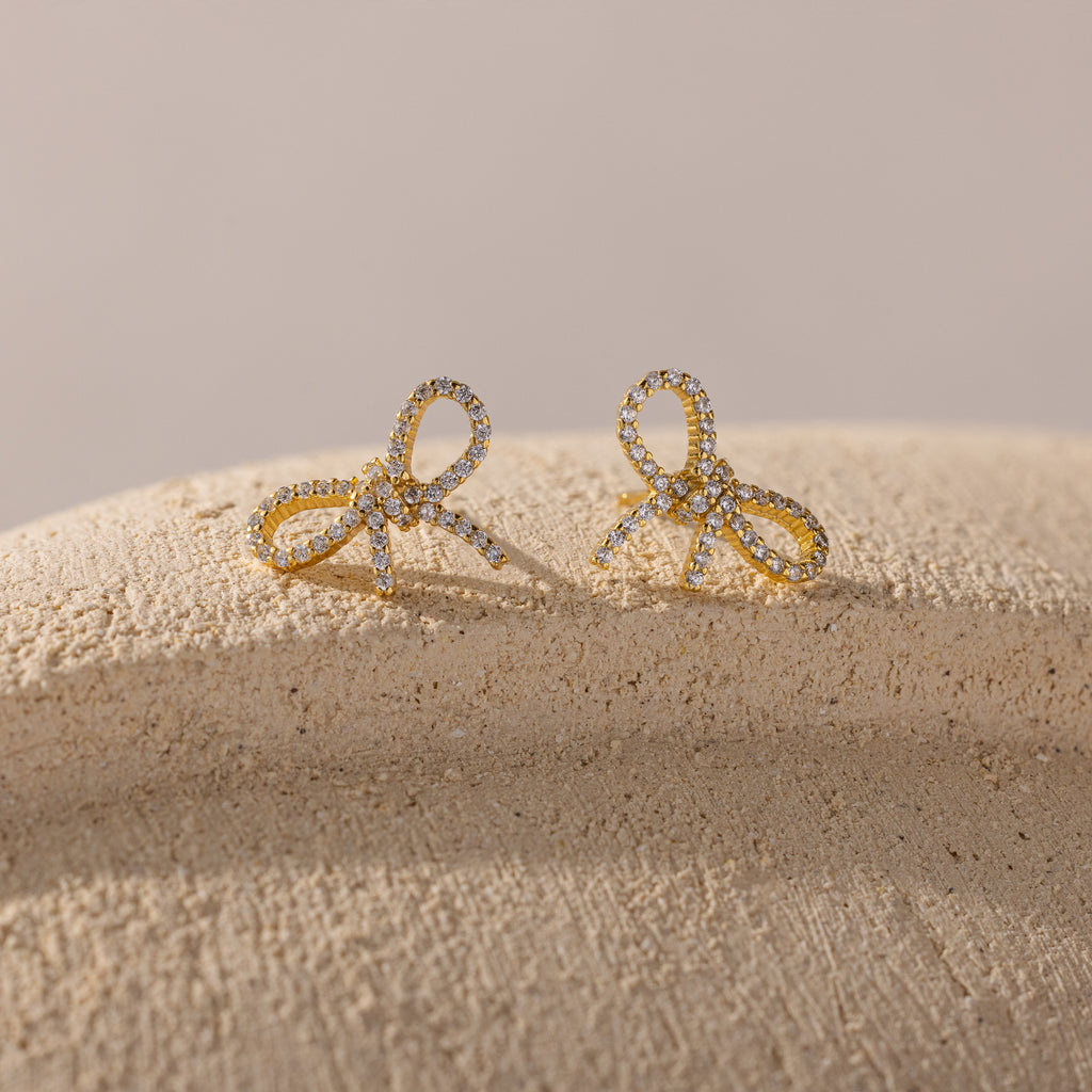 Gold bow-shaped stud earrings encrusted with sparkling cubic zirconia, displayed on a textured neutral surface with soft lighting.