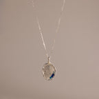 The Organic Fingerprint Multi Birthstone Necklace in Sterling Silver features a textured oval pendant with blue accents, ideal as a personalized keepsake or unique fingerprint necklace, presented on a plain beige background.