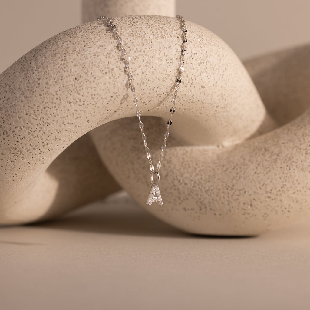 The Dainty Pavé Letter Necklace in Sterling Silver features a sparkling letter A pendant elegantly displayed over a textured, cream abstract sculpture.