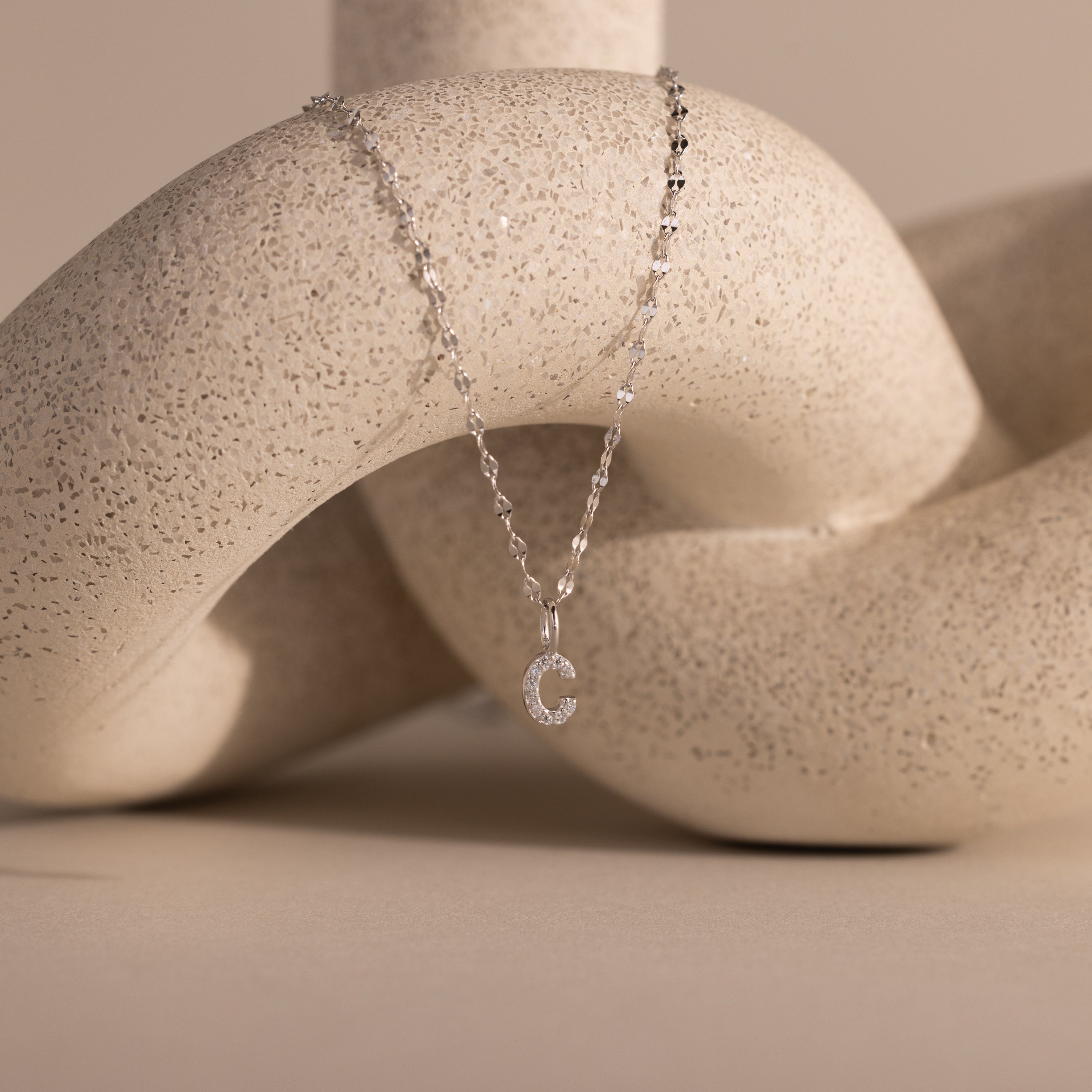 The Dainty Pave Letter Necklace in Sterling Silver, featuring a sparkling letter C pendant, is draped over a textured, abstract beige sculpture.