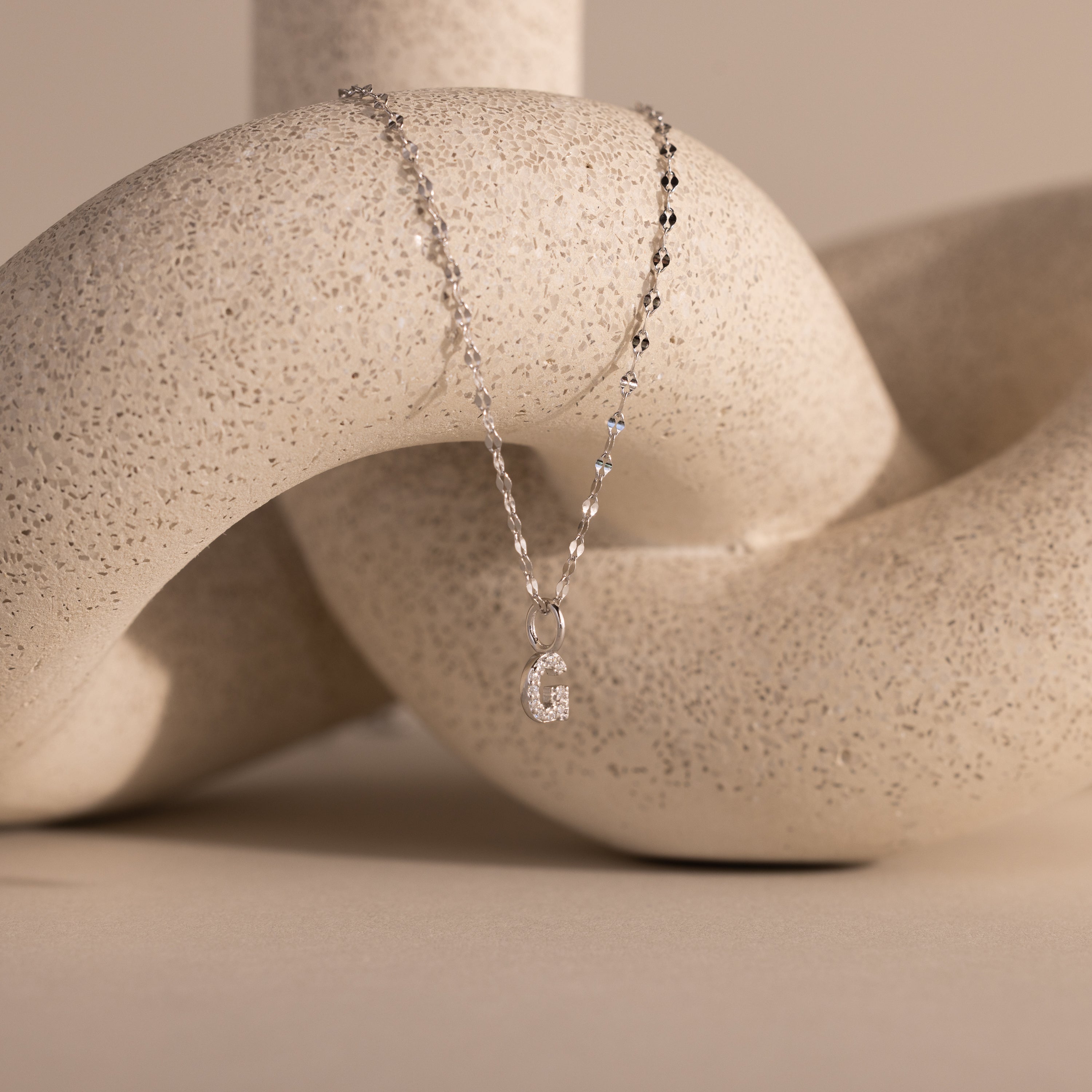 The Dainty Pave Letter Necklace in Sterling Silver, featuring a pavé letter G pendant, drapes elegantly over a textured, abstract beige sculpture.