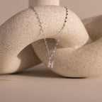 The Dainty Pave Letter Necklace in Sterling Silver features an H pendant and is shown draped over a textured beige abstract sculpture.