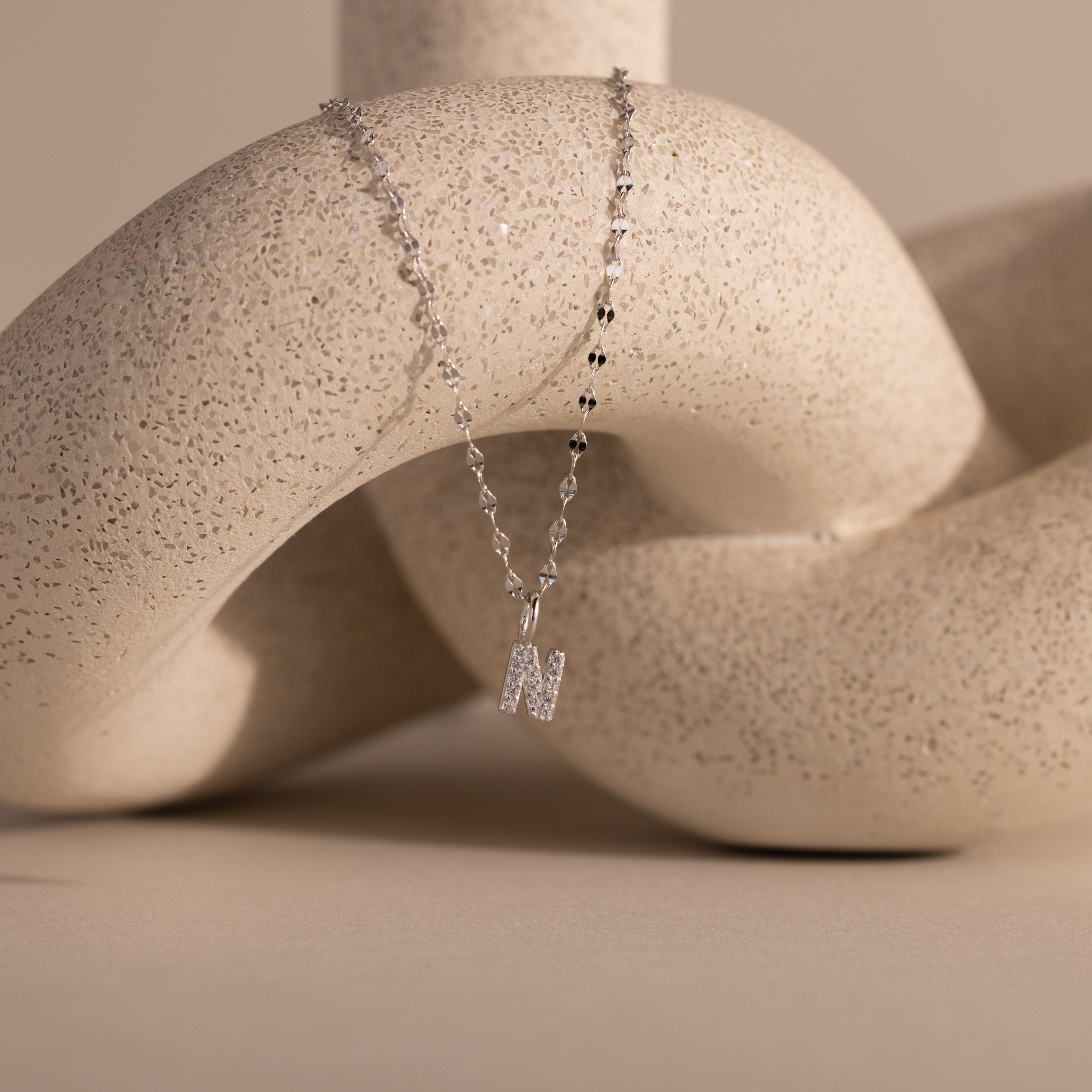 The Dainty Pave Letter Necklace in Sterling Silver, featuring an N pendant with sparkling CZ stones, is showcased draped over a textured, cream-colored abstract sculpture.