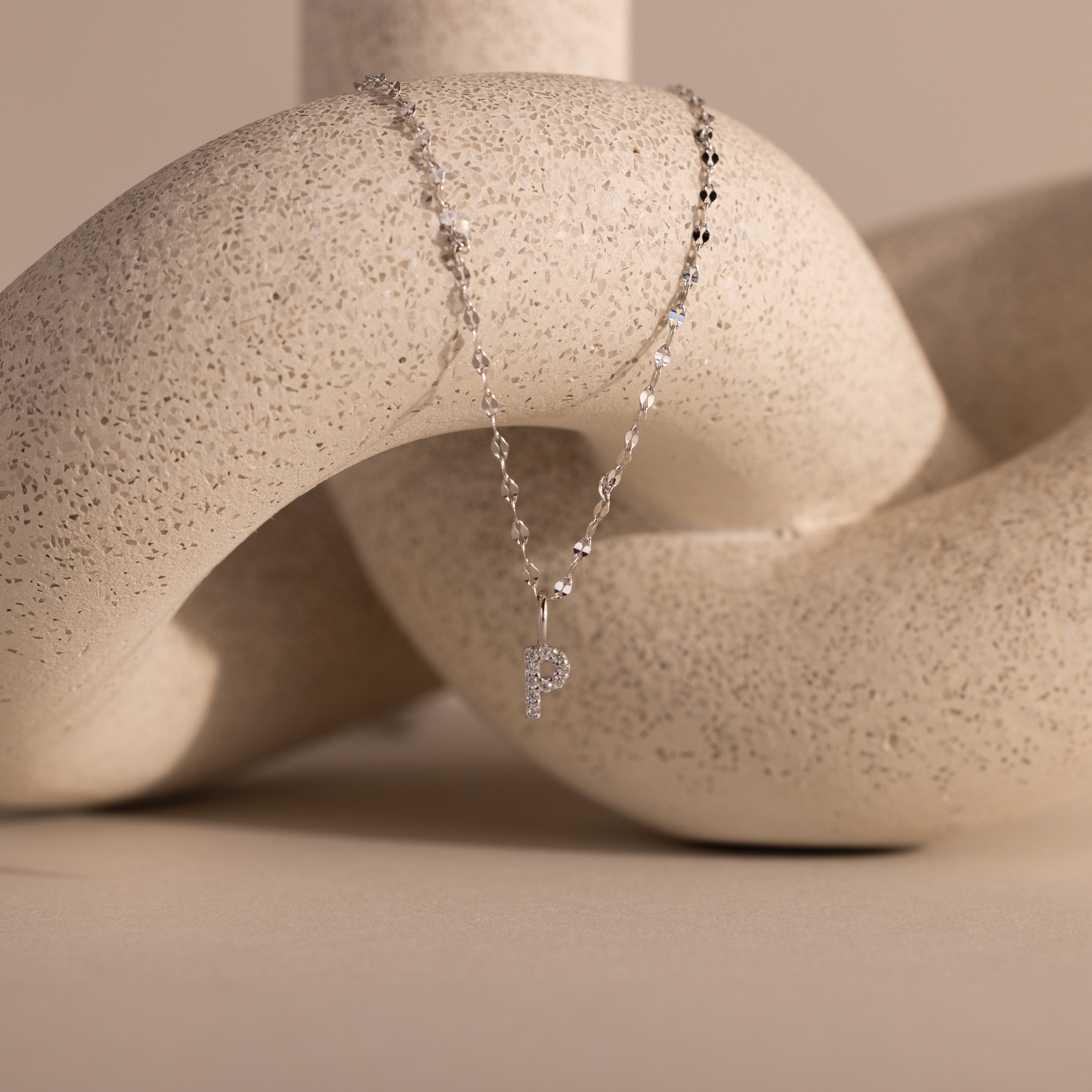 The Dainty Pave Letter Necklace in Sterling Silver features a shimmering pavé P pendant, sparkling like CZ diamonds, elegantly draped over a textured abstract beige sculpture.