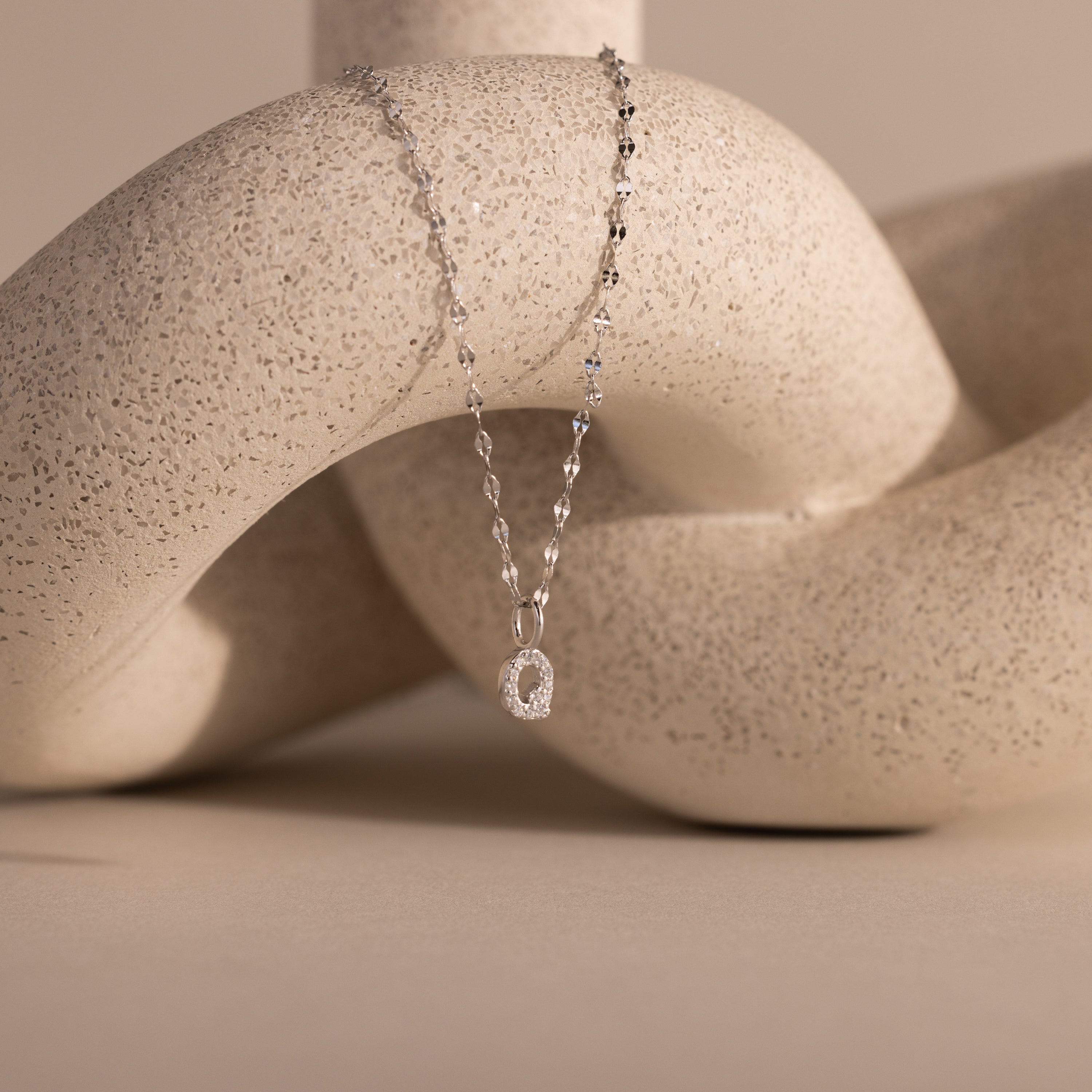 The Dainty Pave Letter Necklace in Sterling Silver features an elegant pendant, beautifully draped over a textured, abstract beige sculpture.