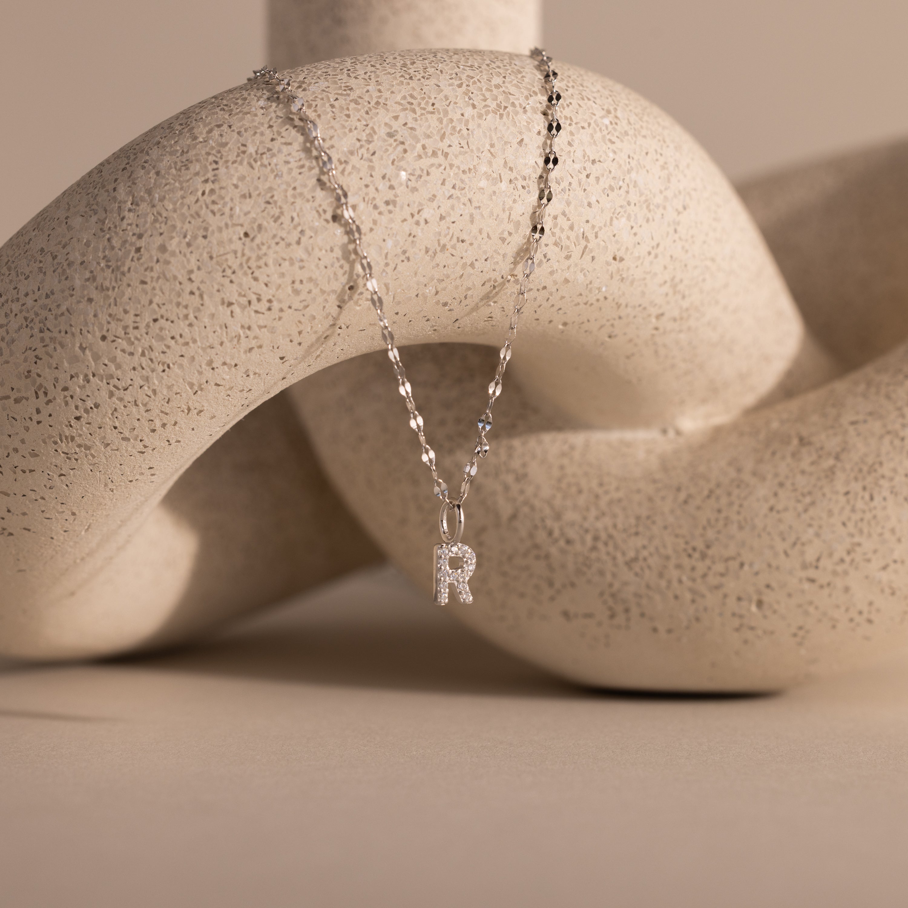 The Dainty Pave Letter Necklace in Sterling Silver features a delicate R pendant adorned with sparkling pavé, draped elegantly over a textured beige sculpture.