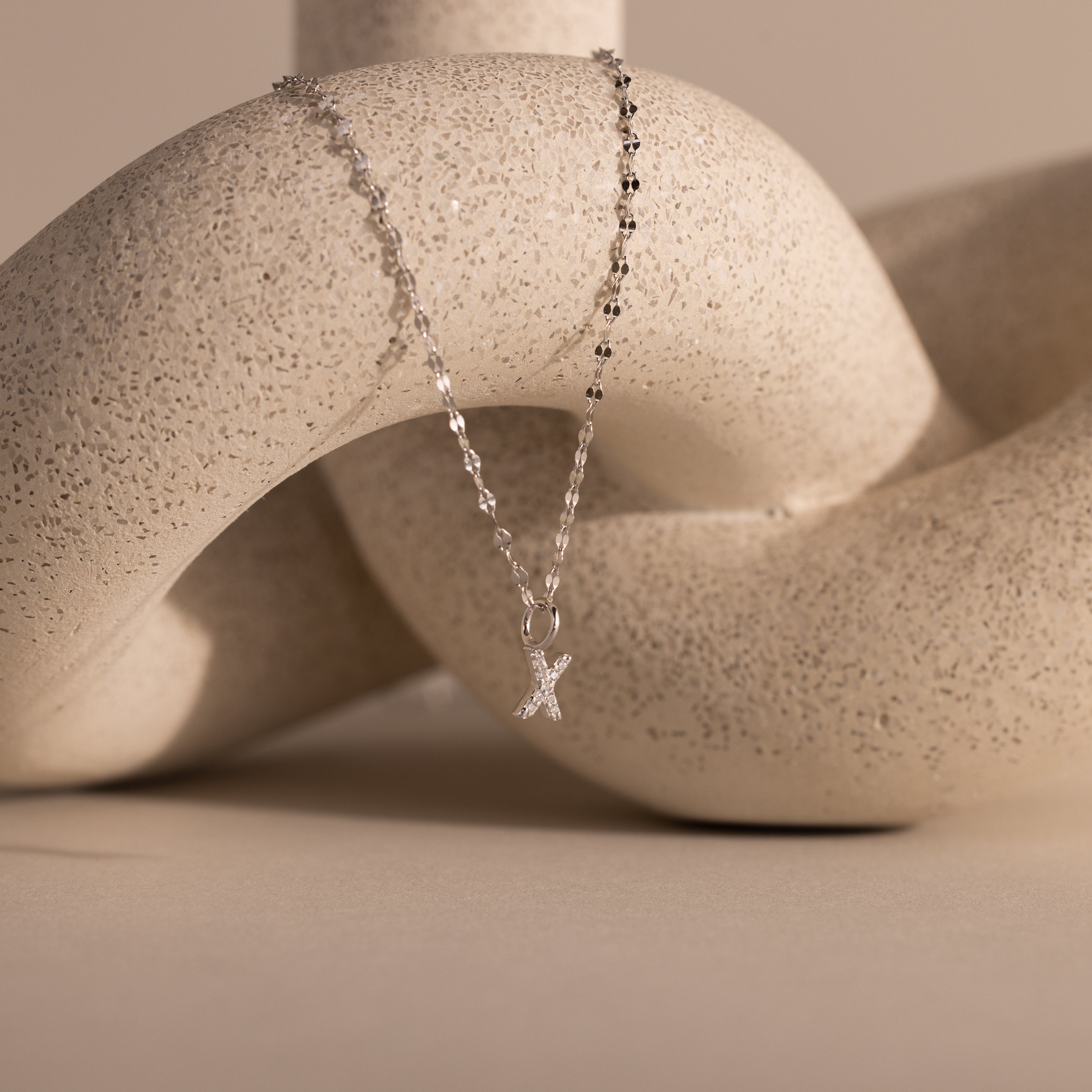 A Dainty Pave Letter Necklace in Sterling Silver drapes elegantly over a textured, abstract beige sculpture.