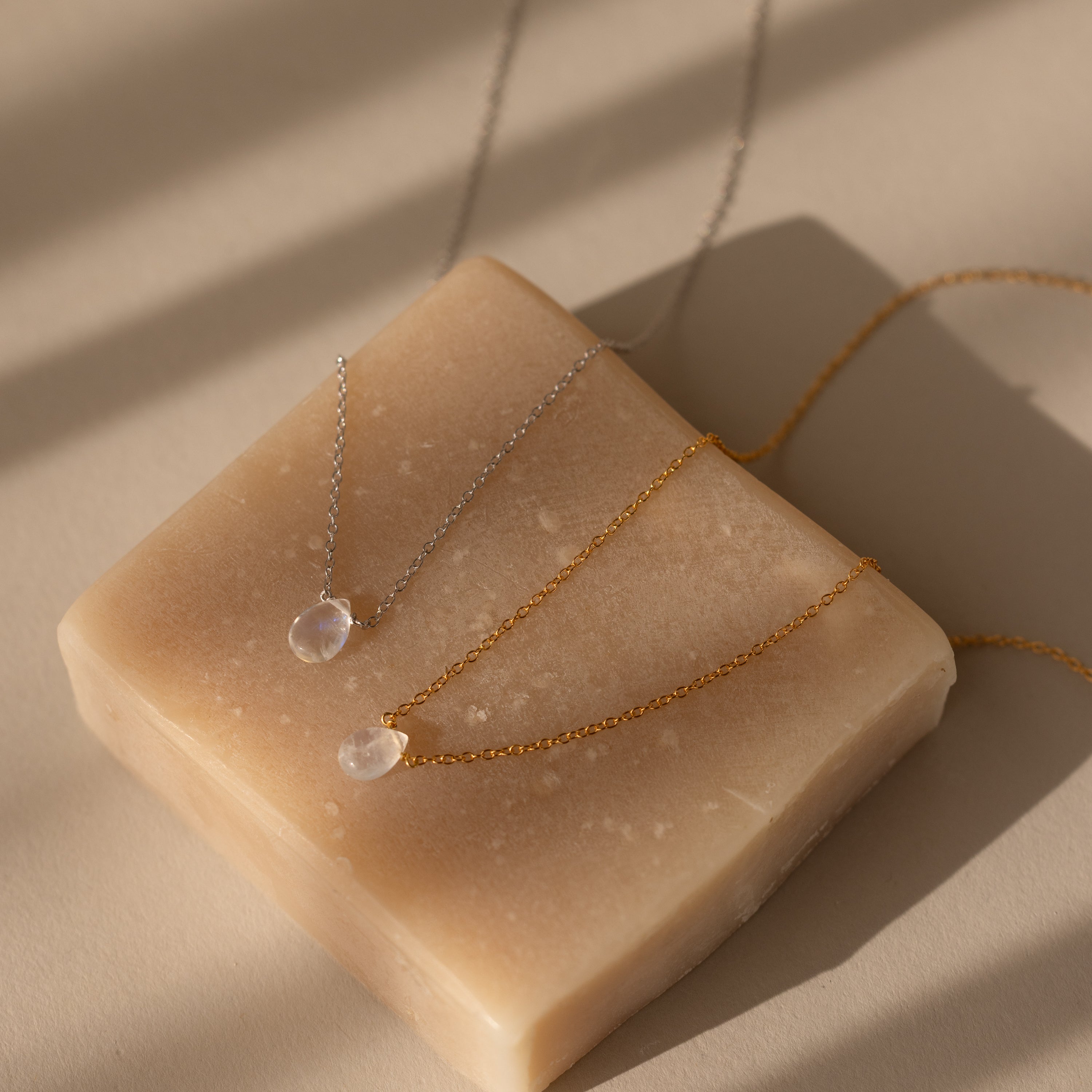 The Teardrop Moonstone Necklace, a delicate minimalist jewelry piece with a pendant, rests on a beige soap bar illuminated by soft, natural light and gentle shadows.