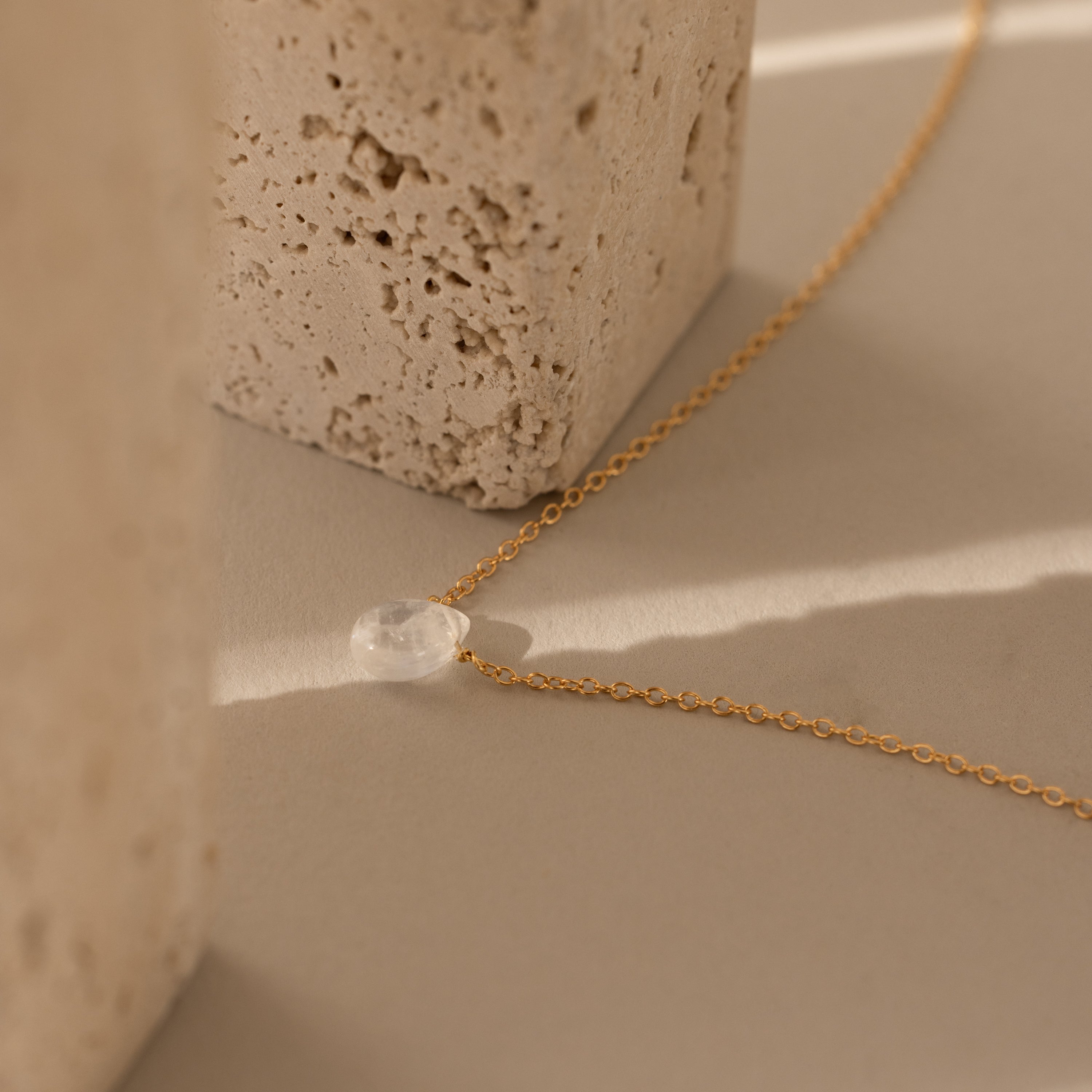 The Teardrop Moonstone Necklace in 18K Gold, featuring a clear stone pendant, rests on a beige surface next to textured stone blocks.