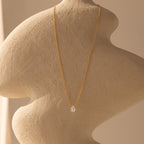 The Teardrop Moonstone Necklace in 18K Gold features a clear moonstone pendant on a gold chain, beautifully displayed on a textured beige abstract sculpture.