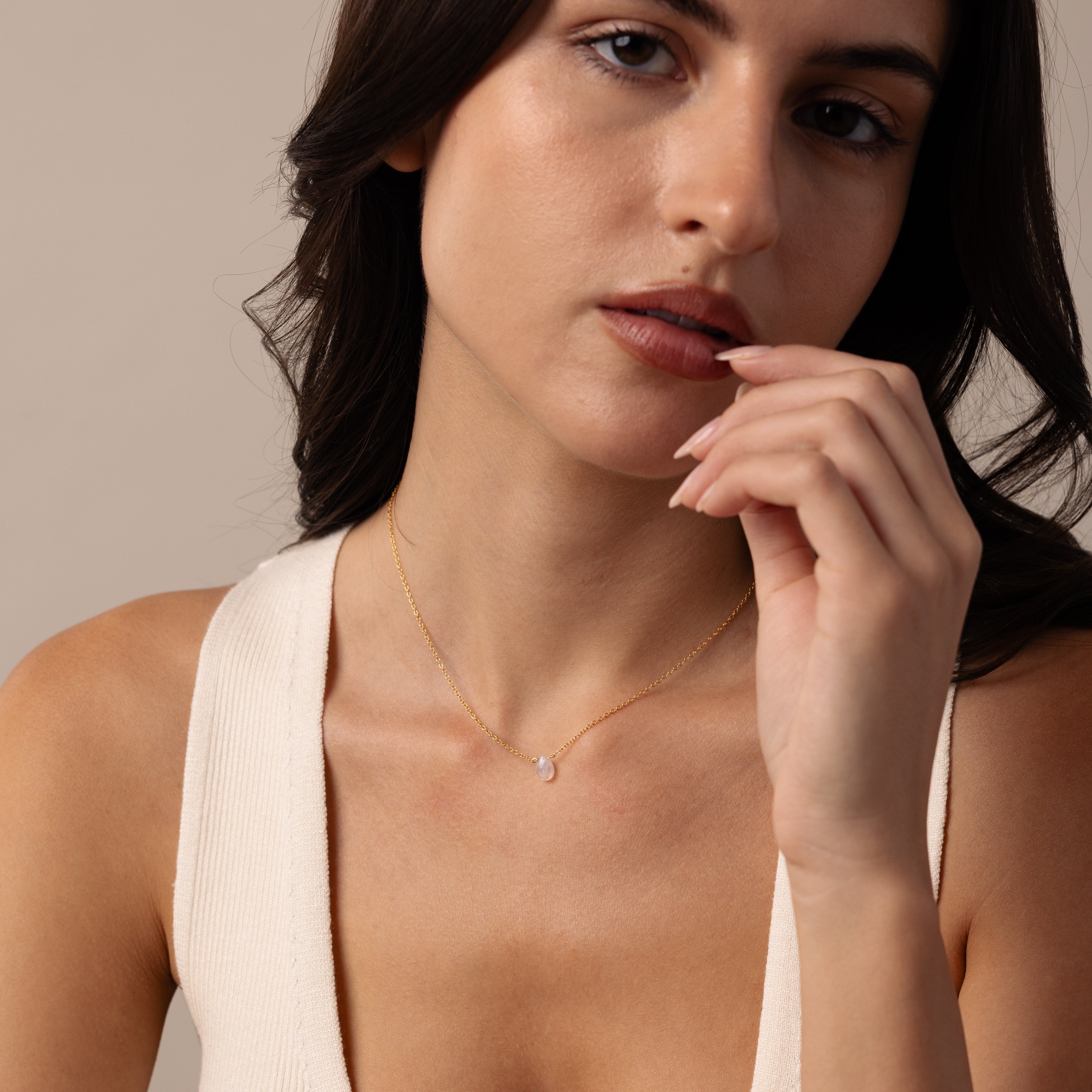 A woman with long dark hair and a white top poses thoughtfully, wearing the Teardrop Moonstone Necklace.