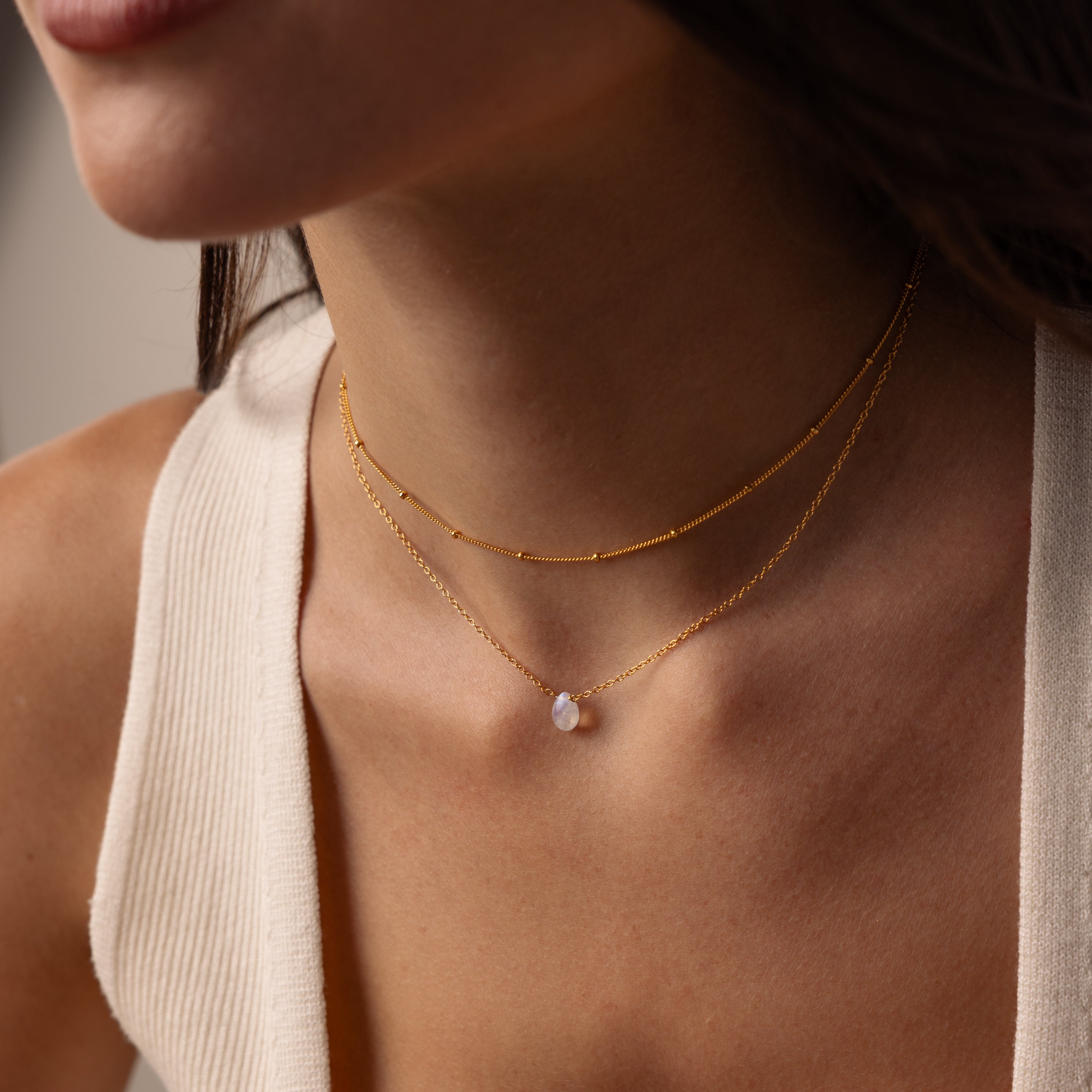 A woman wears the Teardrop Moonstone Necklace layered with another delicate gold necklace over a white ribbed tank top.