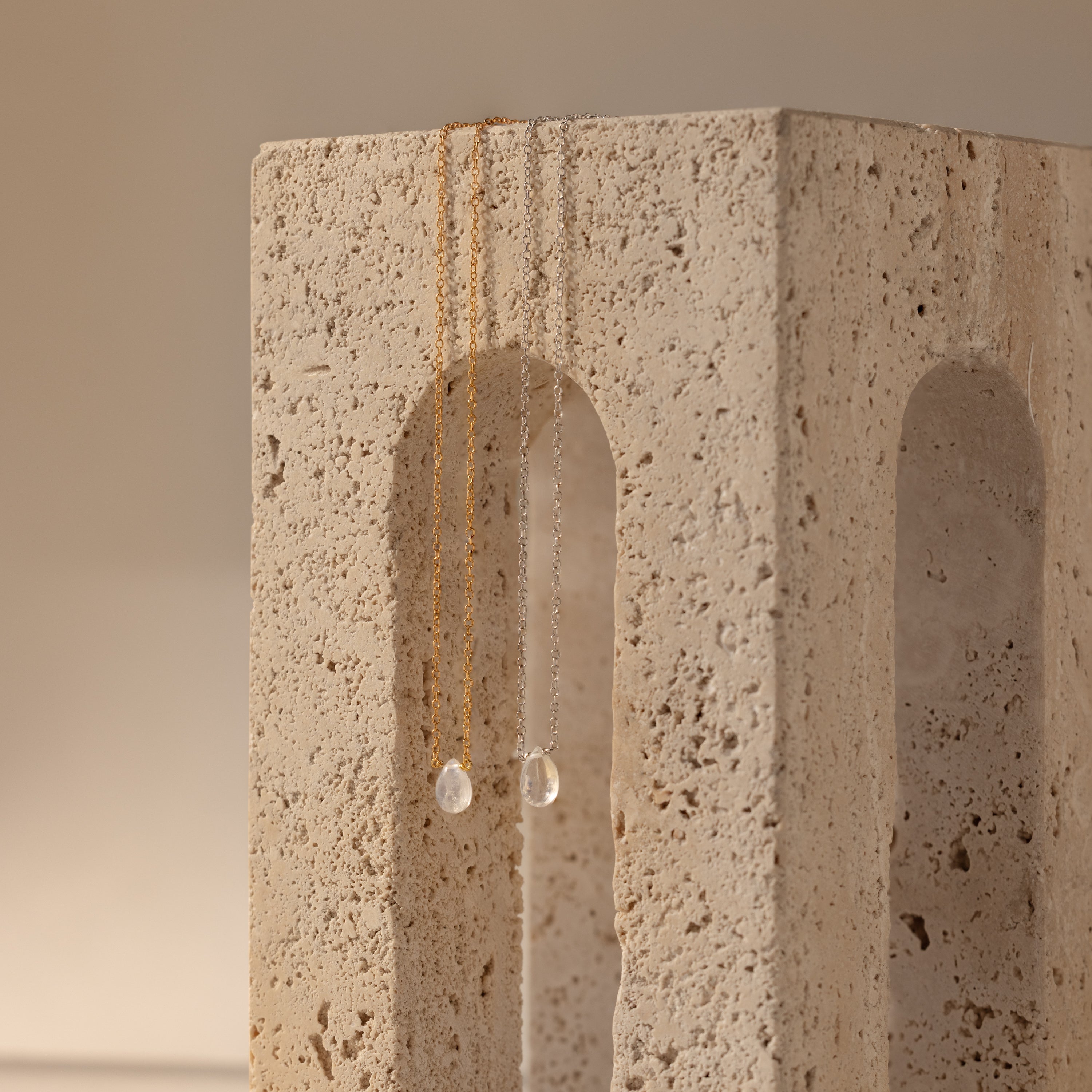 The Teardrop Moonstone Necklace, a minimalist chain adorned with clear teardrop pendants, rests delicately on a textured stone block featuring an arched cutout.