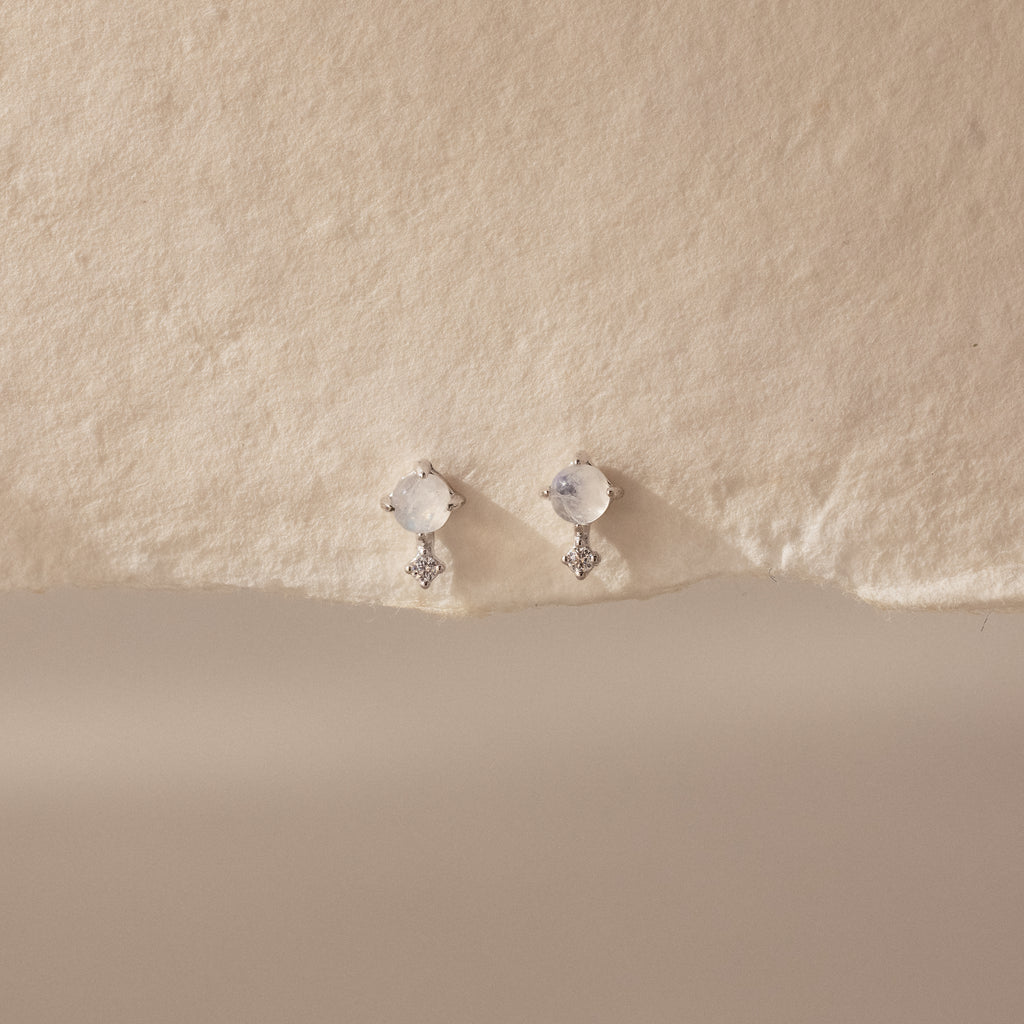 The Moonstone Orbit Studs, featuring round moonstones set in silver, rest on a textured cream surface, bringing a touch of elegance.