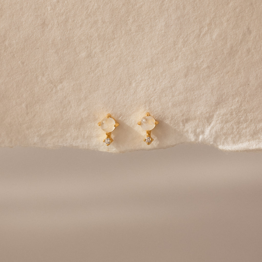 Moonstone Orbit Studs feature translucent moonstones and delicate crystal accents, exuding subtle elegance on textured beige paper. Perfect diamond-accent earrings for any occasion.