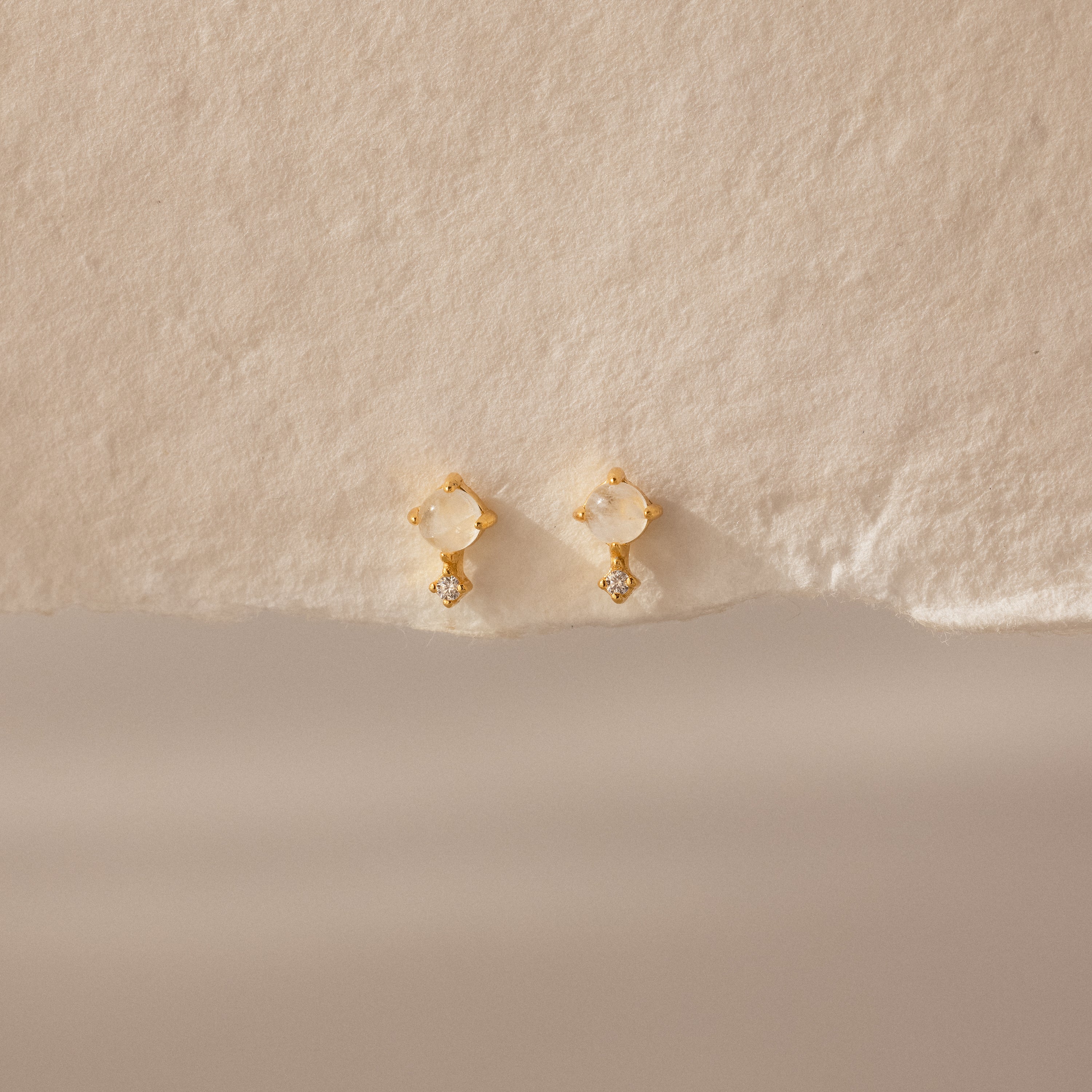 Moonstone Orbit Studs feature translucent moonstones and delicate crystal accents, exuding subtle elegance on textured beige paper. Perfect diamond-accent earrings for any occasion.