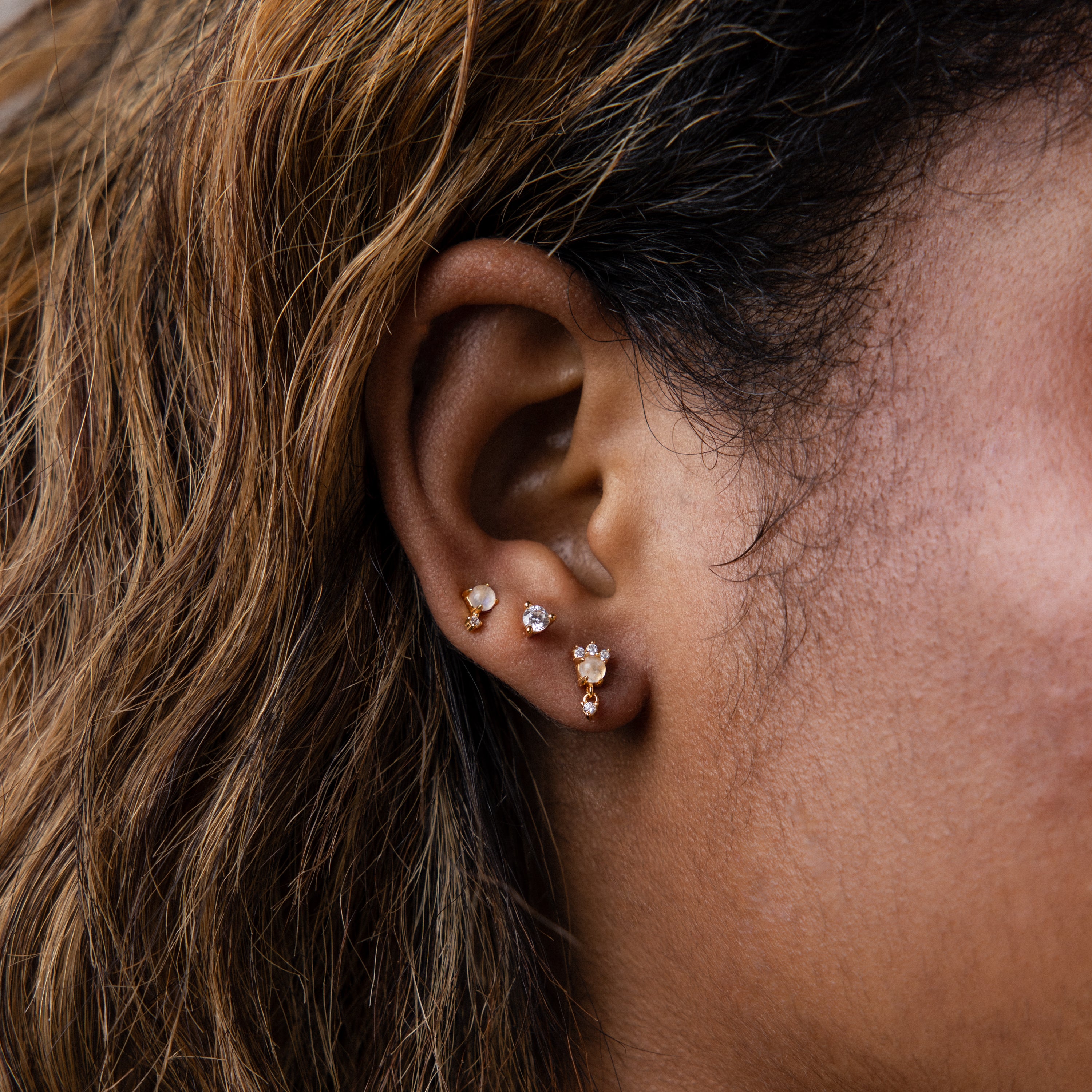 Close-up of a woman's ear with four gold earrings, including diamond accent earrings and Moonstone Orbit Studs in 18K Gold, visible against her wavy brown hair.