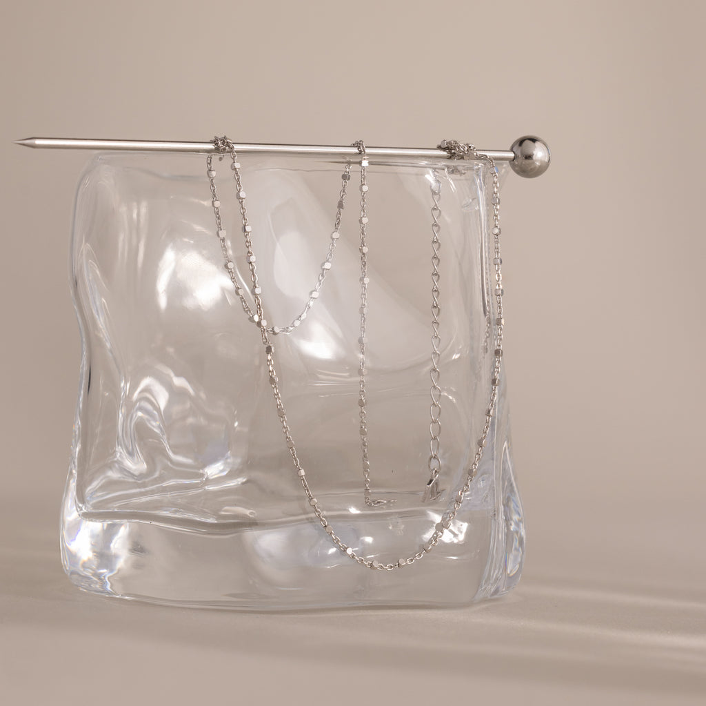 The Dot Station Necklace, a delicate minimalist chain, is draped over a clear wavy glass block with a silver rod resting on top.