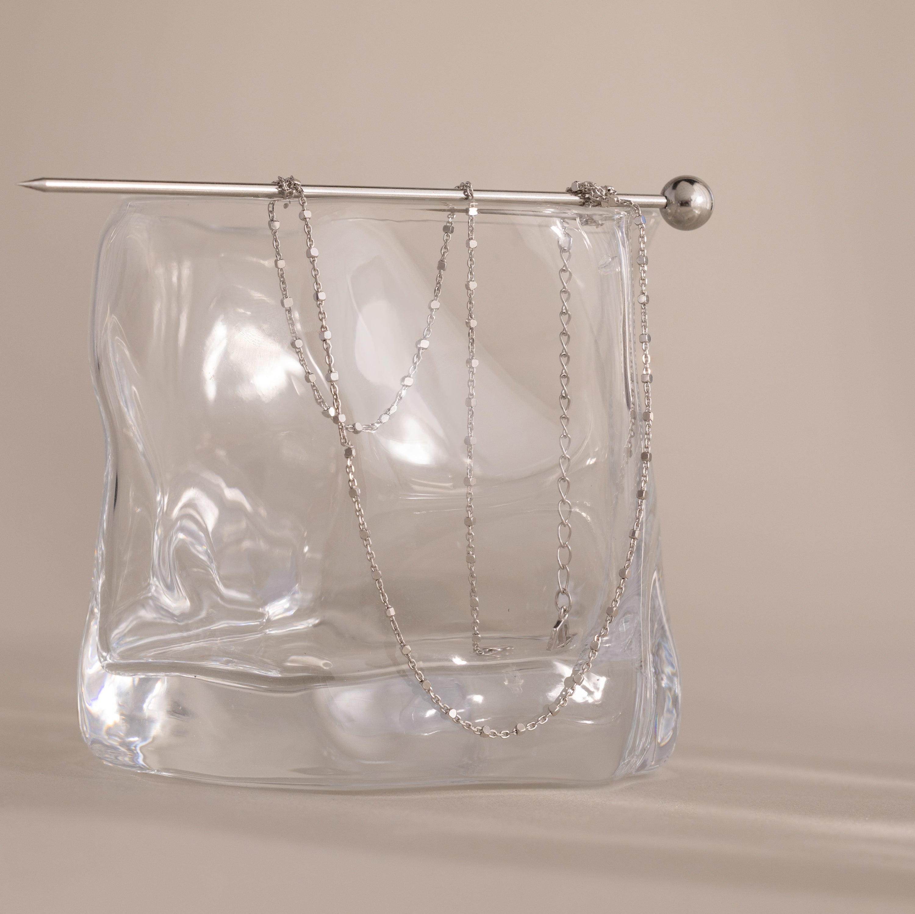 The Dot Station Necklace, a delicate minimalist chain, is draped over a clear wavy glass block with a silver rod resting on top.