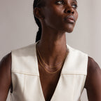 Woman in a sleeveless cream top wears the Woven Chain Necklace layered with a charm necklace, gazing upward with a neutral expression.