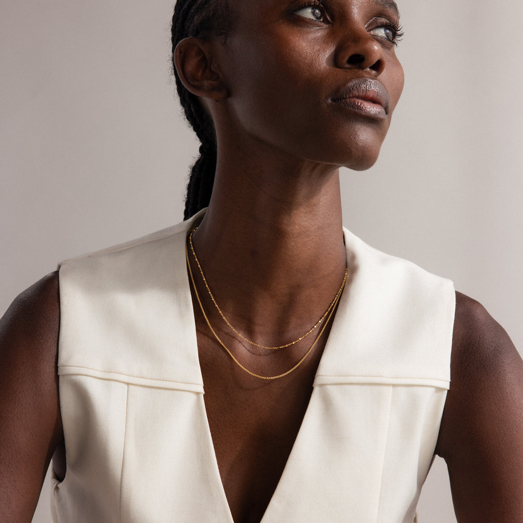 A woman in a cream sleeveless top wears the Dot Station Necklace layered with gold chains, gazing thoughtfully to the side.