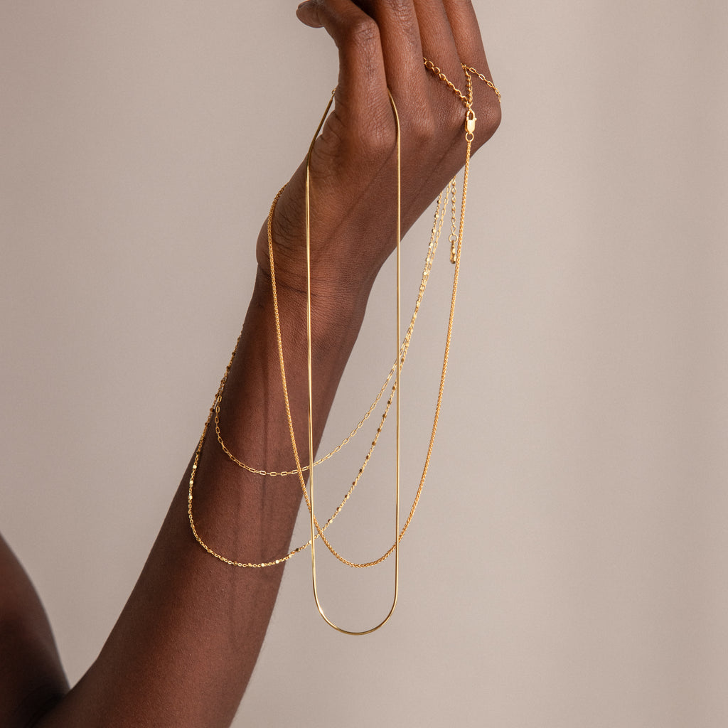 A hand displays the Woven Chain Necklace and other delicate gold necklaces against a neutral background.
