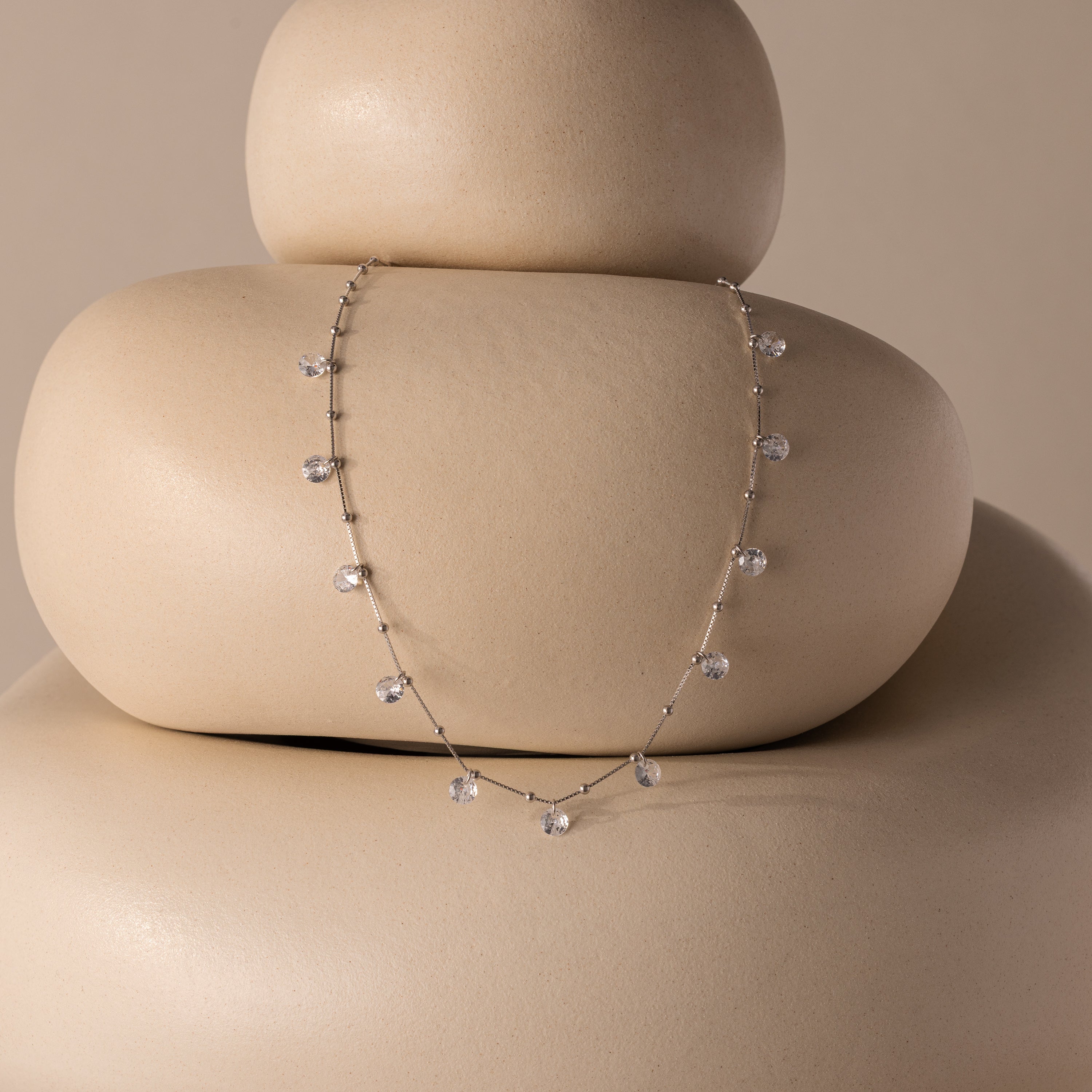 The Satellite Diamond Necklace in Silver features delicate diamond drops and teardrop-shaped crystals elegantly draped over smooth, stacked beige stones.