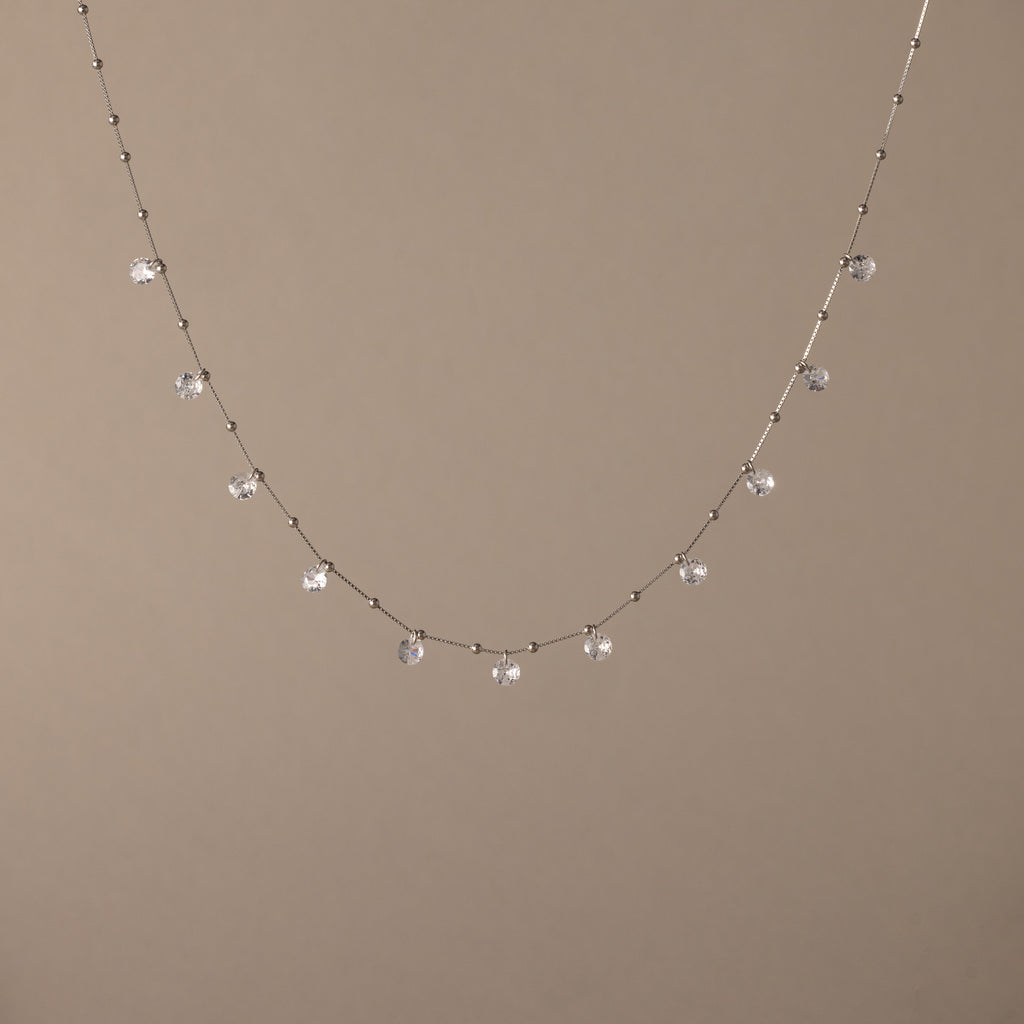 The Satellite Diamond Necklace in Silver features dainty, dangling clear gemstones, reminiscent of a diamond drop station necklace, shown on a beige background.