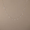 The Satellite Diamond Necklace in Silver features dainty, dangling clear gemstones, reminiscent of a diamond drop station necklace, shown on a beige background.