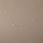 The Satellite Diamond Necklace in Silver features dainty, dangling clear gemstones, reminiscent of a diamond drop station necklace, shown on a beige background.