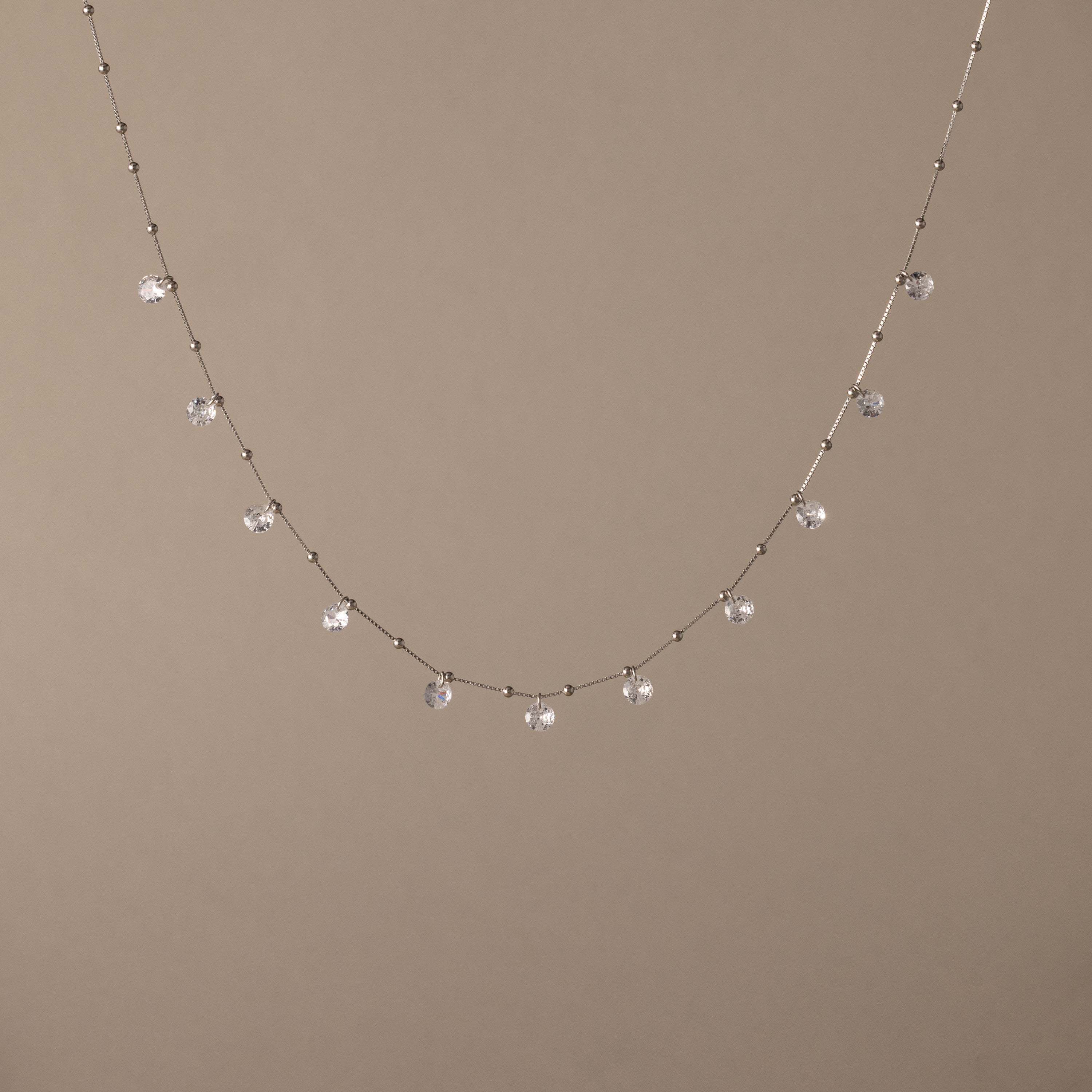 The Satellite Diamond Necklace in Silver features dainty, dangling clear gemstones, reminiscent of a diamond drop station necklace, shown on a beige background.