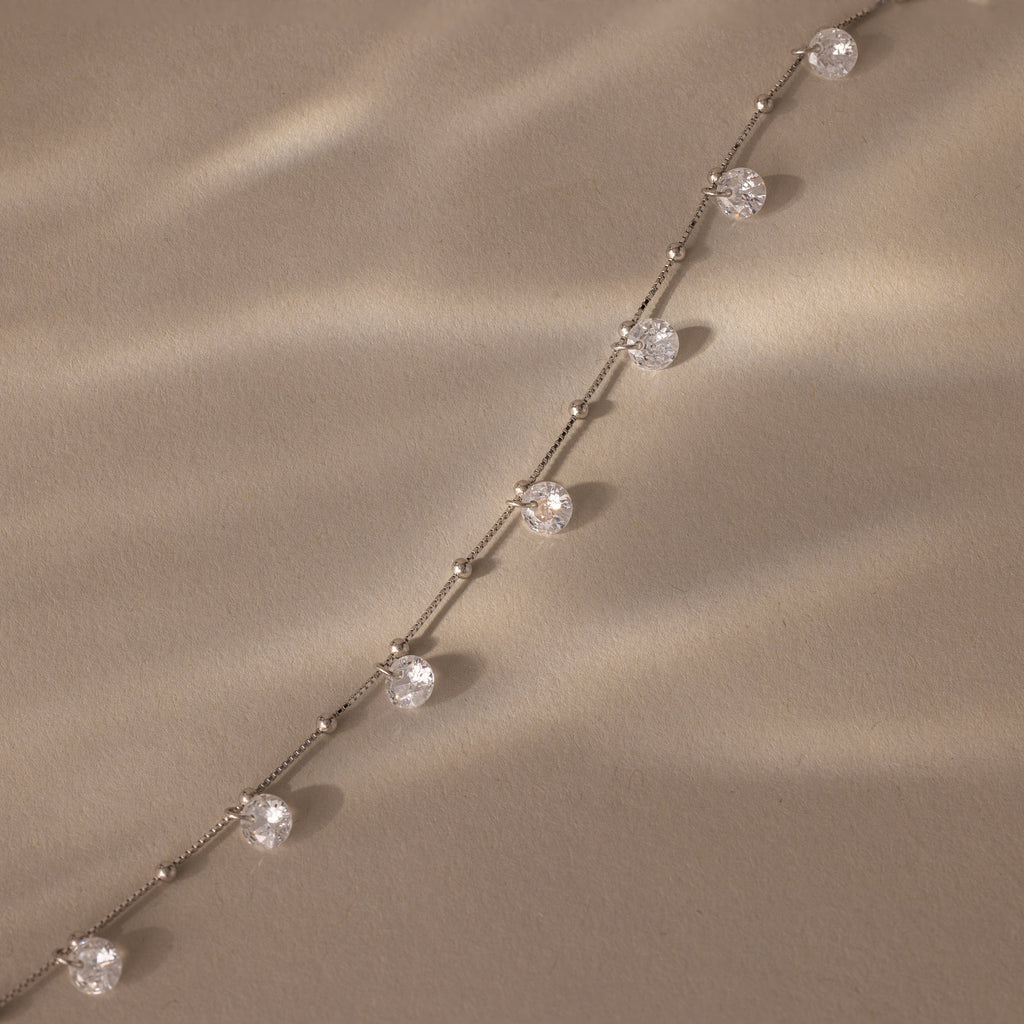 The Satellite Diamond Necklace in Silver, featuring evenly spaced round diamonds, rests on a textured beige surface bathed in soft light.