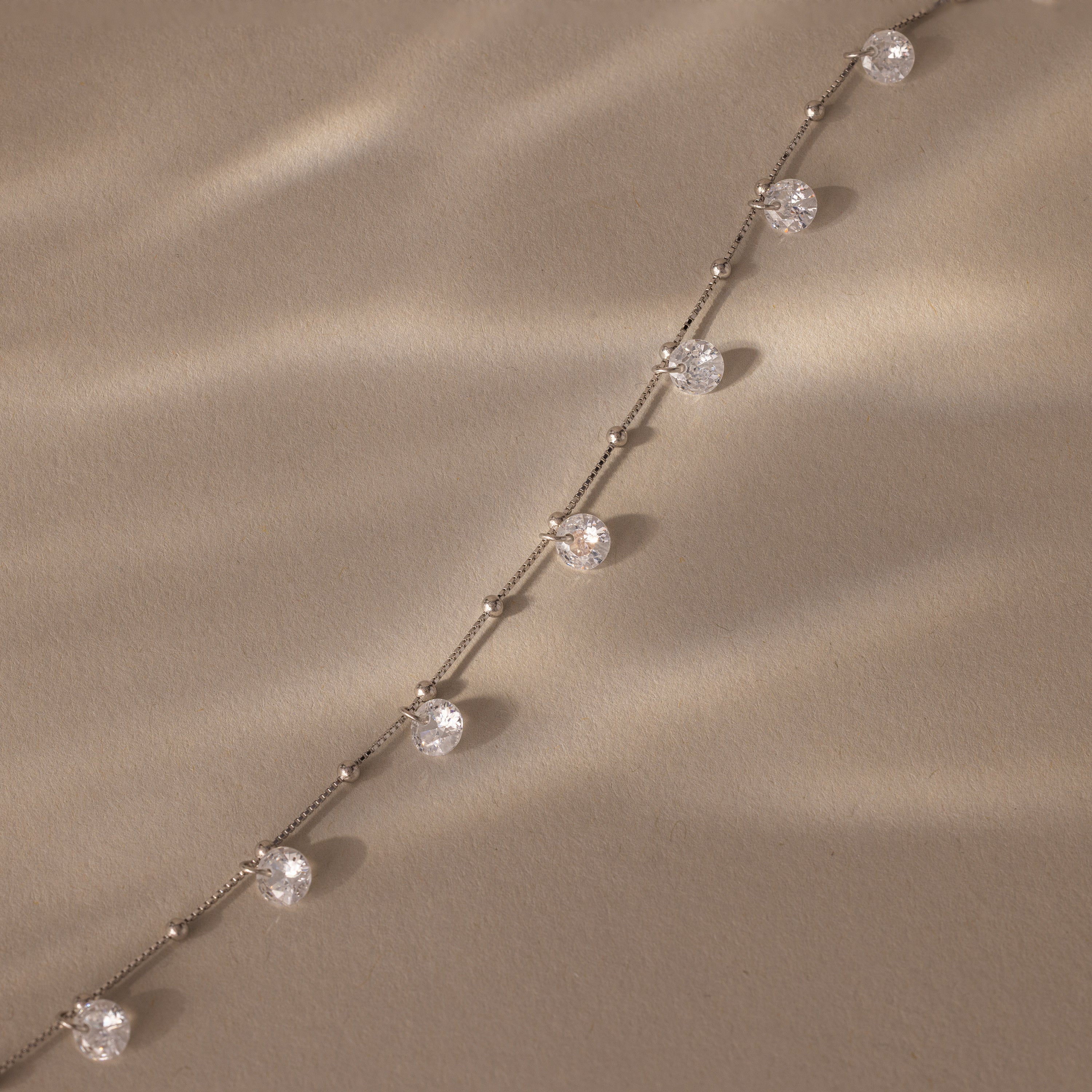 The Satellite Diamond Necklace in Silver, featuring evenly spaced round diamonds, rests on a textured beige surface bathed in soft light.
