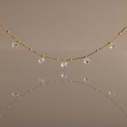 The Satellite Diamond Necklace in 18K Gold features delicate, small clear crystal drops inspired by a classic diamond design, beautifully reflected on a smooth, neutral-colored surface.