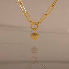 The Paperclip Heart Charm Necklace in 18K Gold features a gold paperclip chain and a heart pendant, elegantly reflected on a smooth surface against a neutral background.