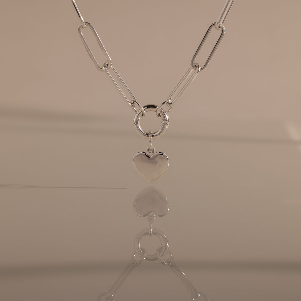The Paperclip Heart Charm Necklace in Silver is displayed on a smooth surface, its heart pendant reflected against a neutral background.