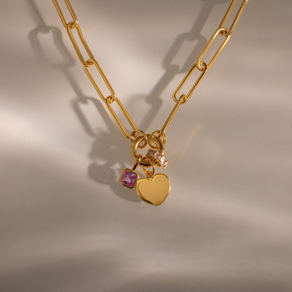 The Paperclip Heart Charm Necklace features a gold paperclip chain with a heart charm and two gemstone charms, showcased on a neutral background with soft shadows.