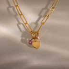 The Paperclip Heart Charm Necklace features a gold paperclip chain with a heart charm and two gemstone charms, showcased on a neutral background with soft shadows.
