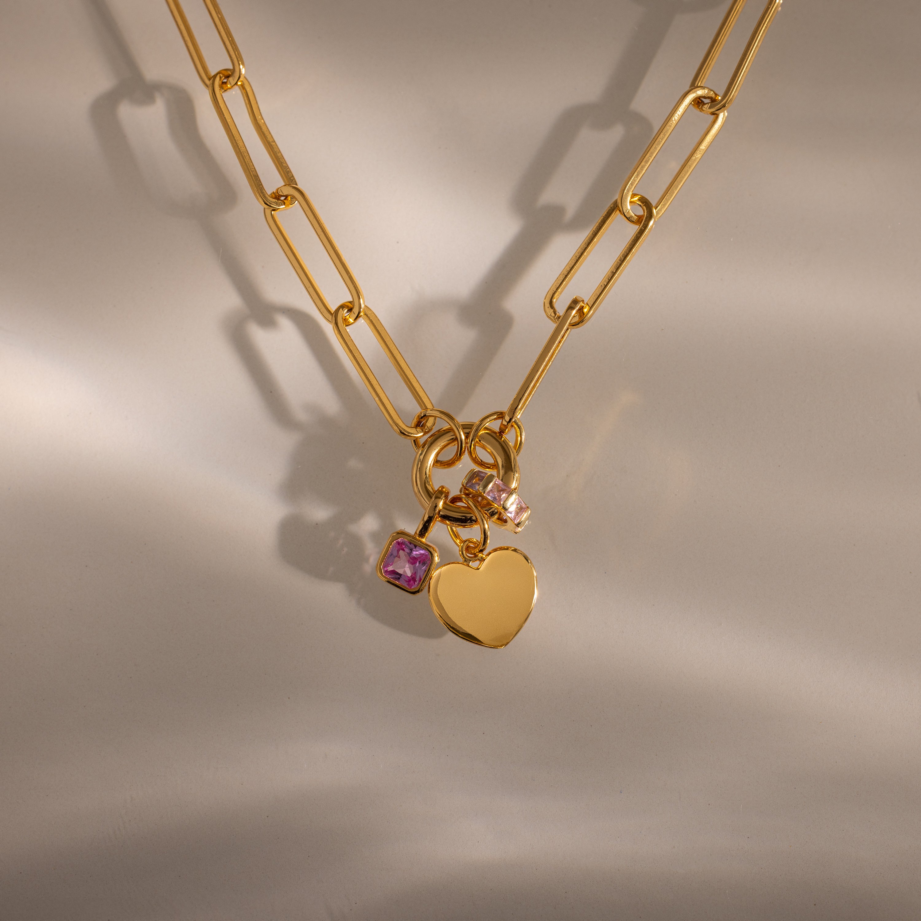 The Paperclip Heart Charm Necklace features a gold paperclip chain with a heart charm and two gemstone charms, showcased on a neutral background with soft shadows.