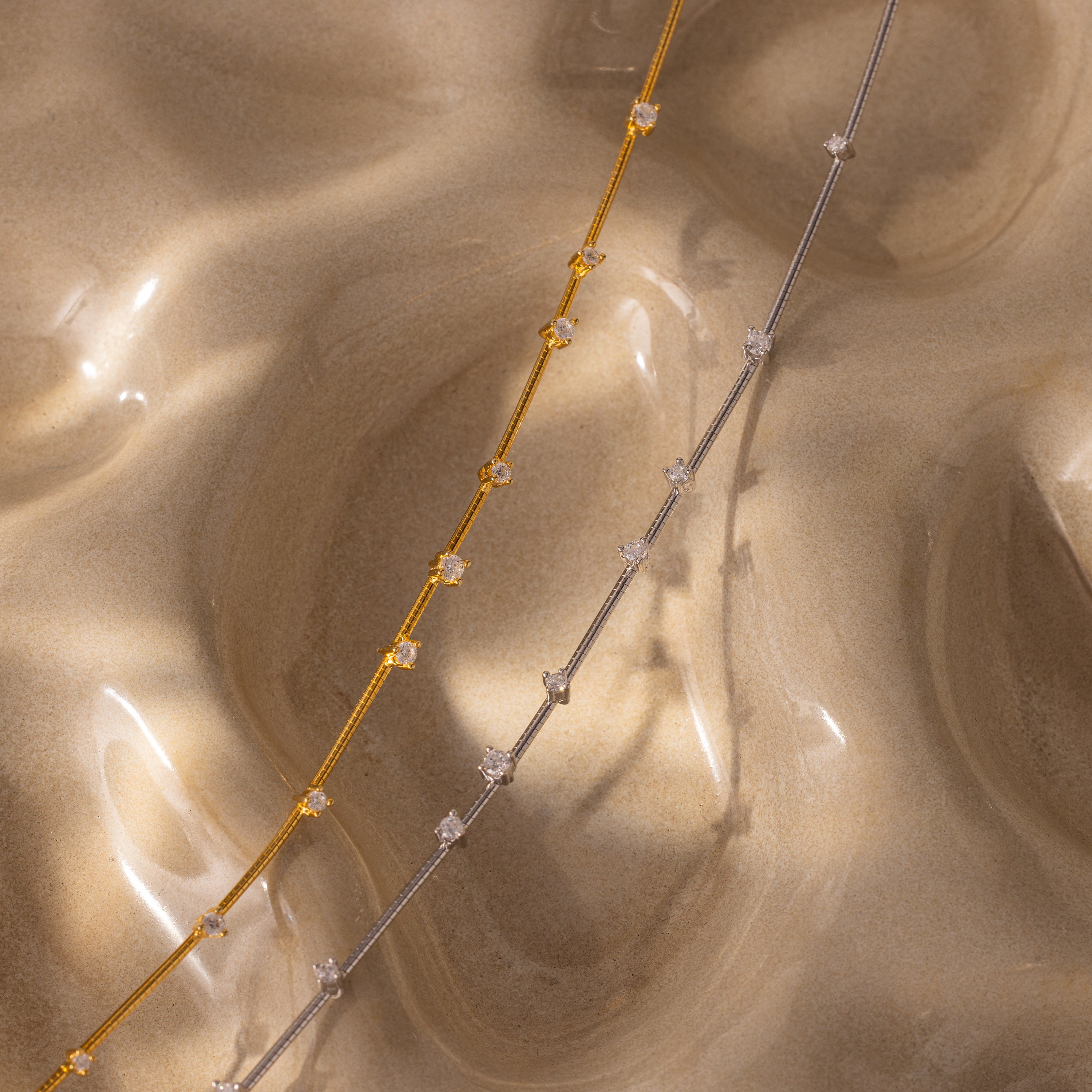 The Sprinkle Diamond Necklace, a delicate chain adorned with small gemstones, rests on a wavy, sand-textured beige surface—perfect for adding a touch of everyday brilliance to any look.