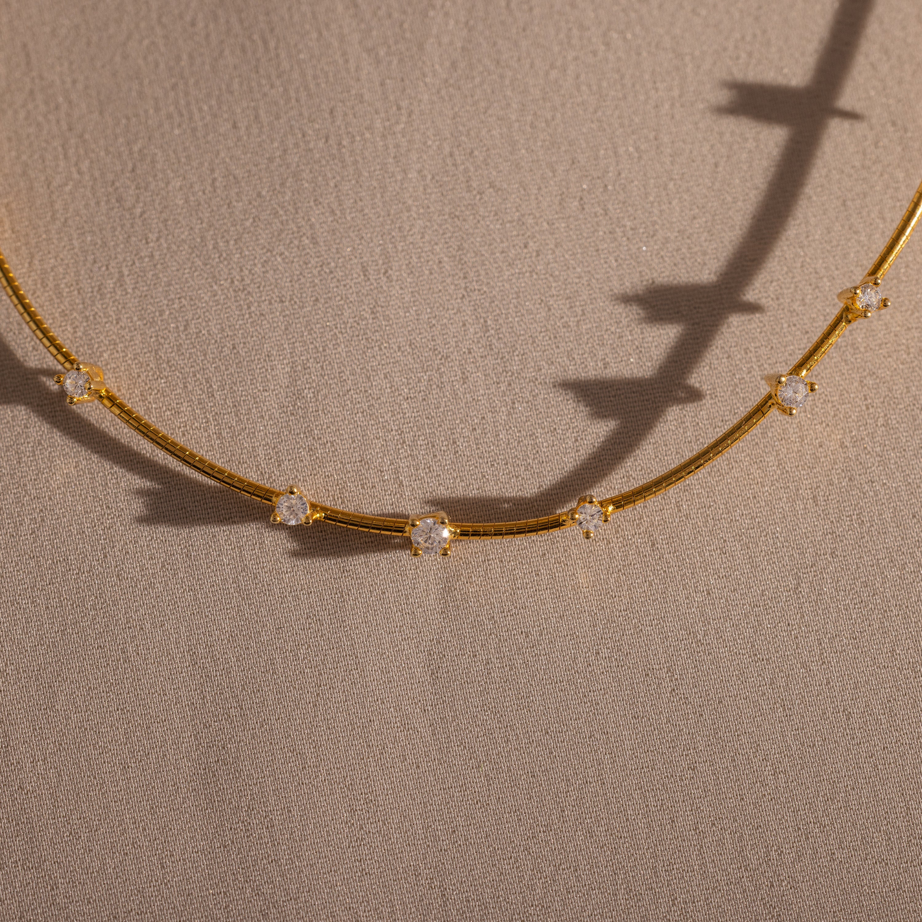 The Sprinkle Diamond Necklace in 18K Gold features a delicate chain adorned with evenly spaced sparkling stones, shown resting on a textured beige surface.
