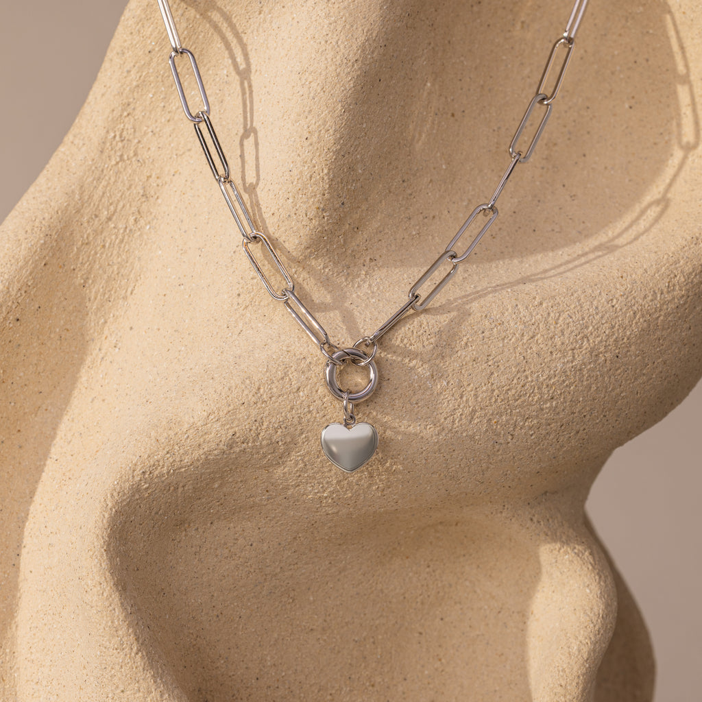 The Paperclip Heart Charm Necklace in Silver features a heart pendant, elegantly displayed on a smooth, beige, abstract sculpture.