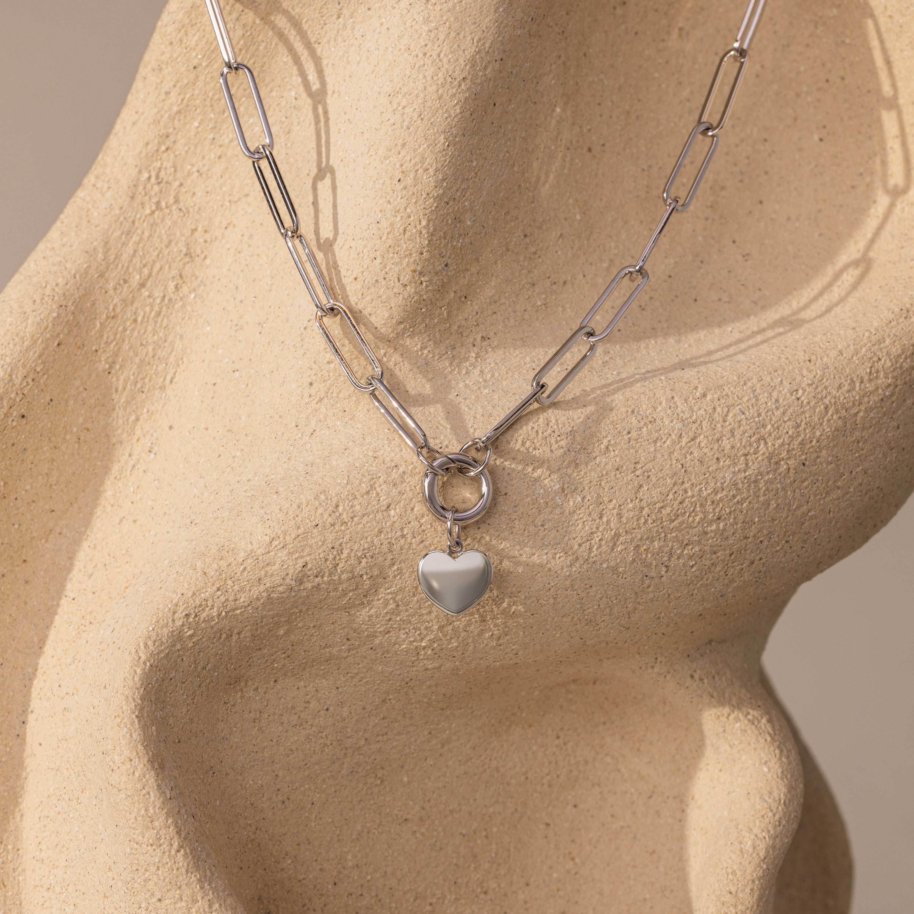 The Paperclip Heart Charm Necklace in Silver features a heart pendant, elegantly displayed on a smooth, beige, abstract sculpture.