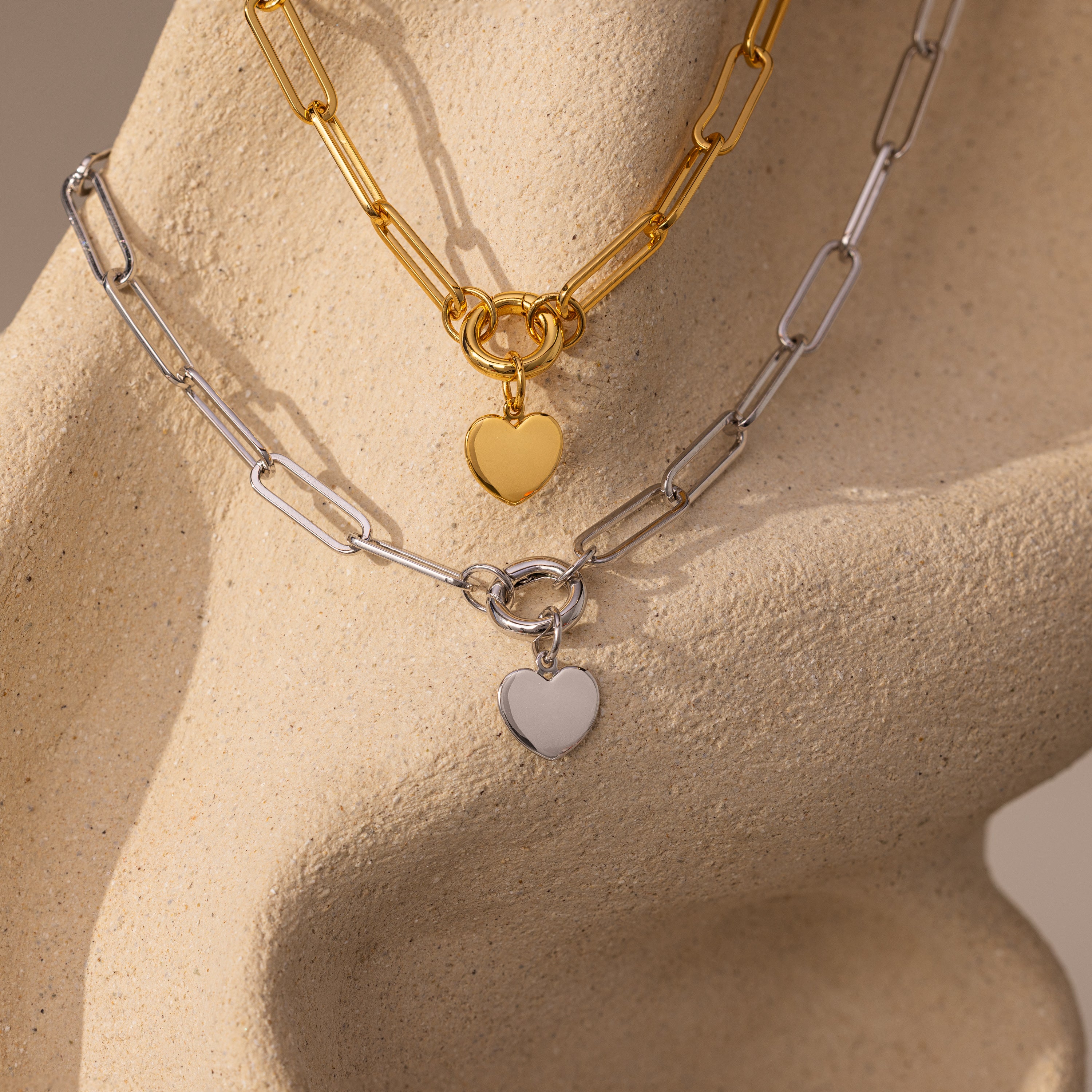 The Paperclip Heart Charm Necklace, featuring gold and silver chain styles with heart pendants, is displayed on a textured beige sculpture.