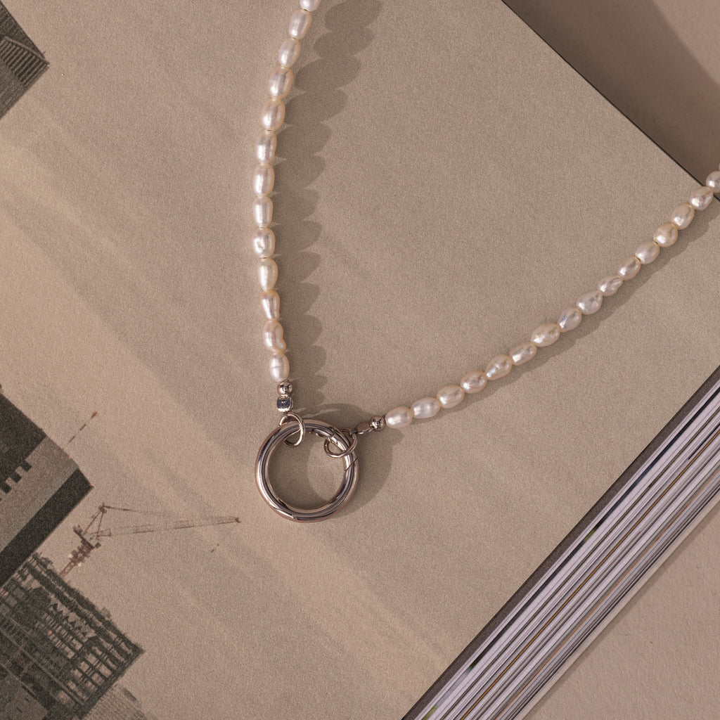 The Pearl Chain Clasp Necklace features modern pearls and a silver ring clasp, beautifully displayed on an open book with architectural images—a timeless addition to any jewelry collection.