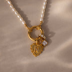 The Pearl Chain Clasp Necklace features a gold heart pendant with pearls and gemstone charms, showcased on a beige surface—a standout addition to any jewelry collection.